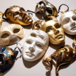 Mehrere abgelegte Masken auf heller Fläche – Symbol für gesellschaftliche Rollen, Täuschung und das, was sichtbar wird, wenn Masken fallen.
